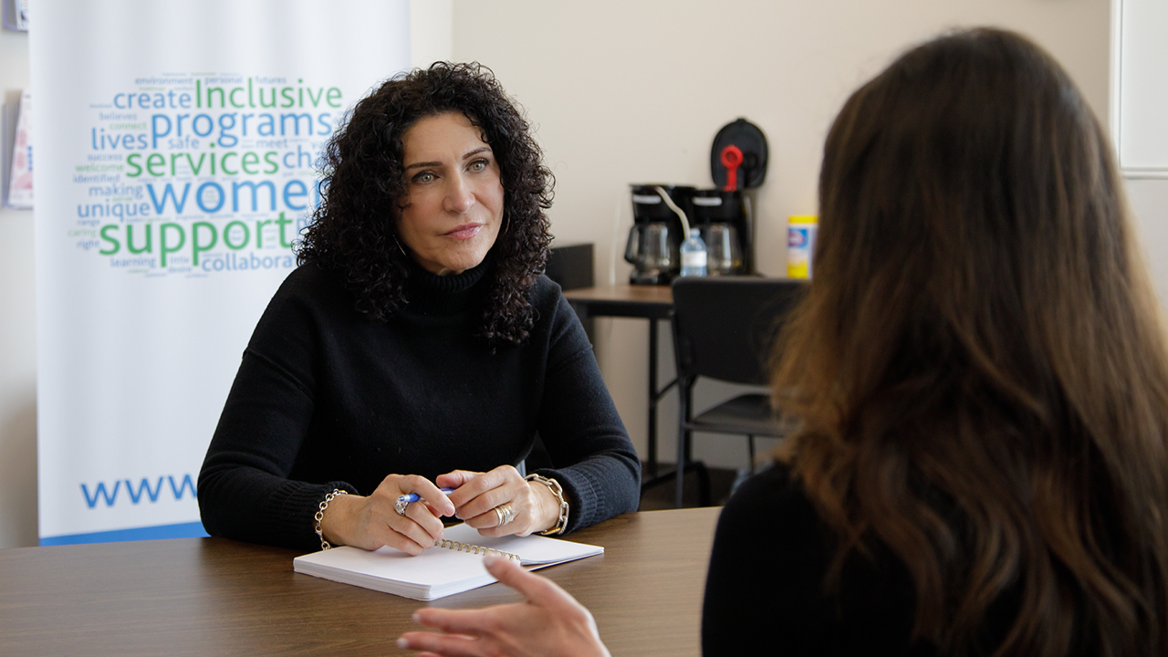 Piera, a light-skinned woman with dark curly hair and wearing a black turtleneck, sits at a desk listening intently to the woman sitting across from her At Women's Centre of York Region
