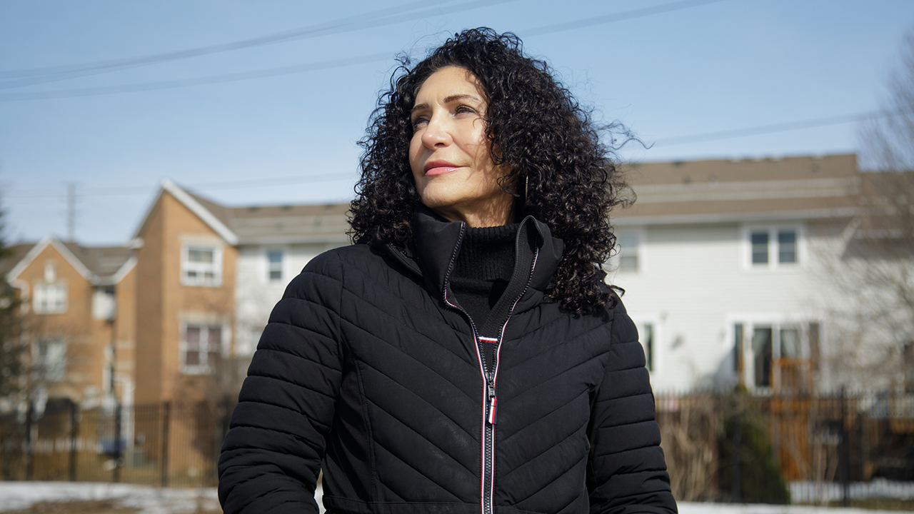 Piera, a light-skinned woman with dark curly hair and wearing a black turtleneck and black winter coat. She looks off into the distance.