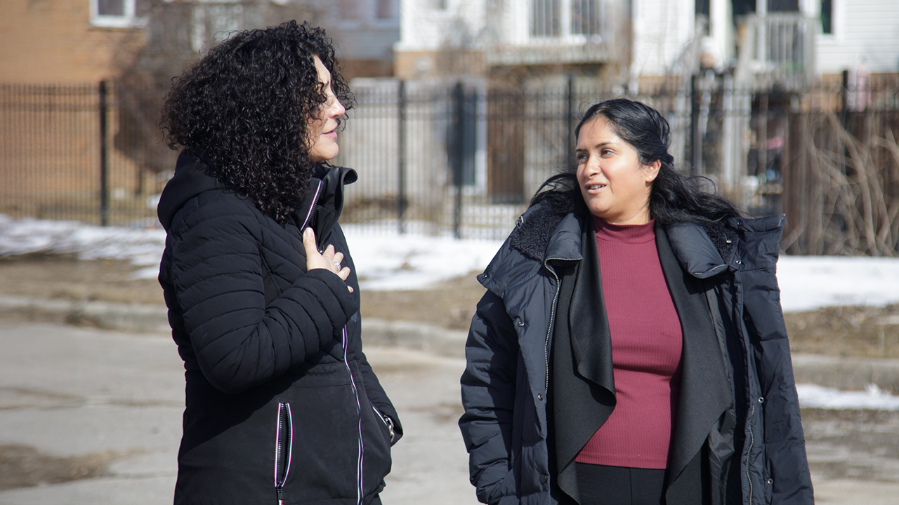 Piera and Nivedita from Women's Centre of York Region walk down a street wearing heavy coats. Snow partly covers the grass in the background.