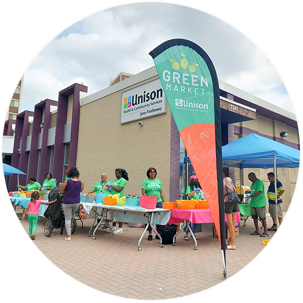 People in green shirts stand behind tables set up outside the front entrance of the United Way-supported Jane Street Community Hub. 
