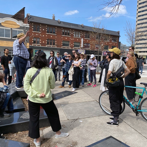 Participants on a United Way neighbourhood tour gather in a semi-circle on a sidewalk, listening to a speaker.