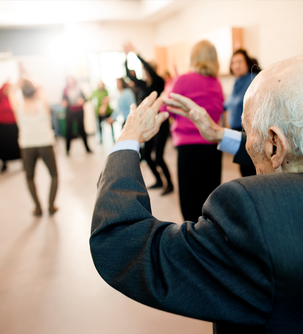 A senior, seen from behind, raises their hands above their head in a gesture, with others in the background doing the same.