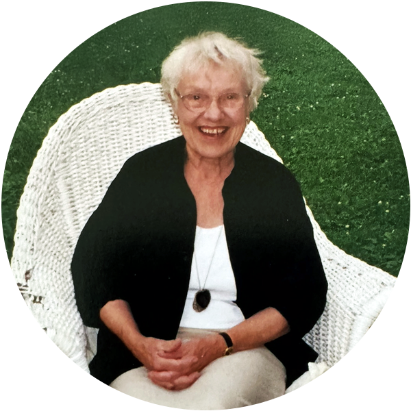 A woman with white hair and wearing a white shirt, black cardigan and khaki pants sits in a white chair on green grass. She is laughing and looking into the camera.