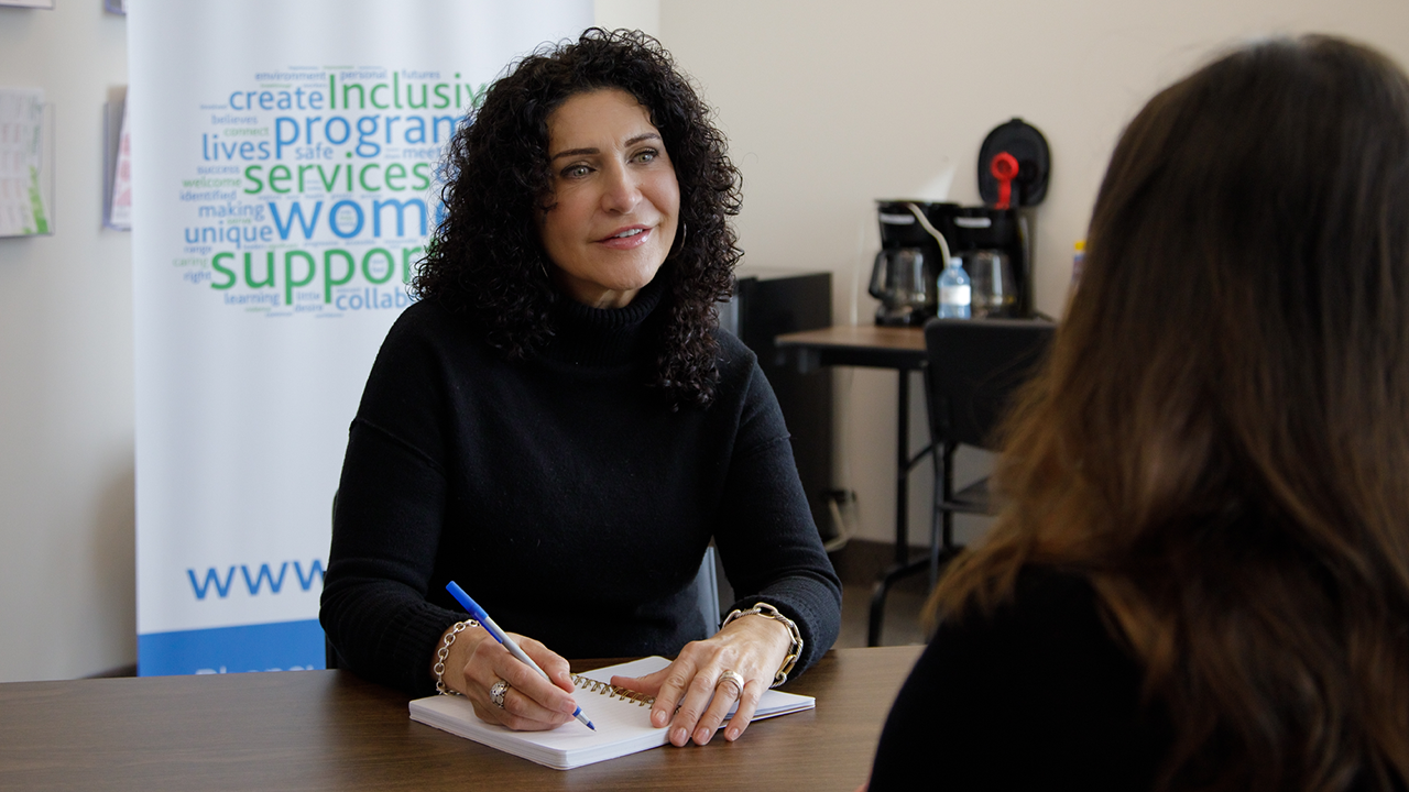 Piera, a light-skinned woman with dark curly hair and wearing a black turtleneck, sits at a desk listening intently to the woman sitting across from her At Women's Centre of York Region
