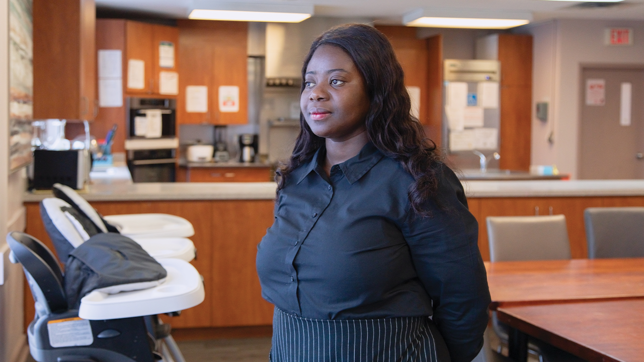 Francisca, a Black woman, is dressed in a black button down top and wears a black stripped apron while standing in the kitchen at the Embrave Agency to End Violence shelter.