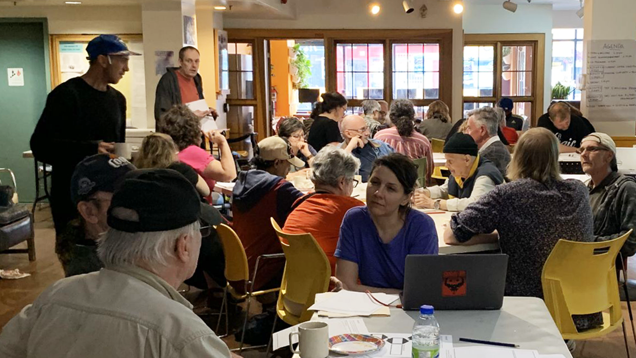 At the United Way-supported Parkdale Activity Recreation Centre, a woman sits at a laptop listening attentively to an older man. Several others are seated at tables in the background, engaged in conversation.