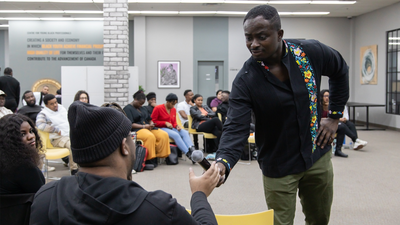 A packed audience watches as a Black man presenting hands a microphone to someone in the audience at CEE Centre for Young Black Professionals