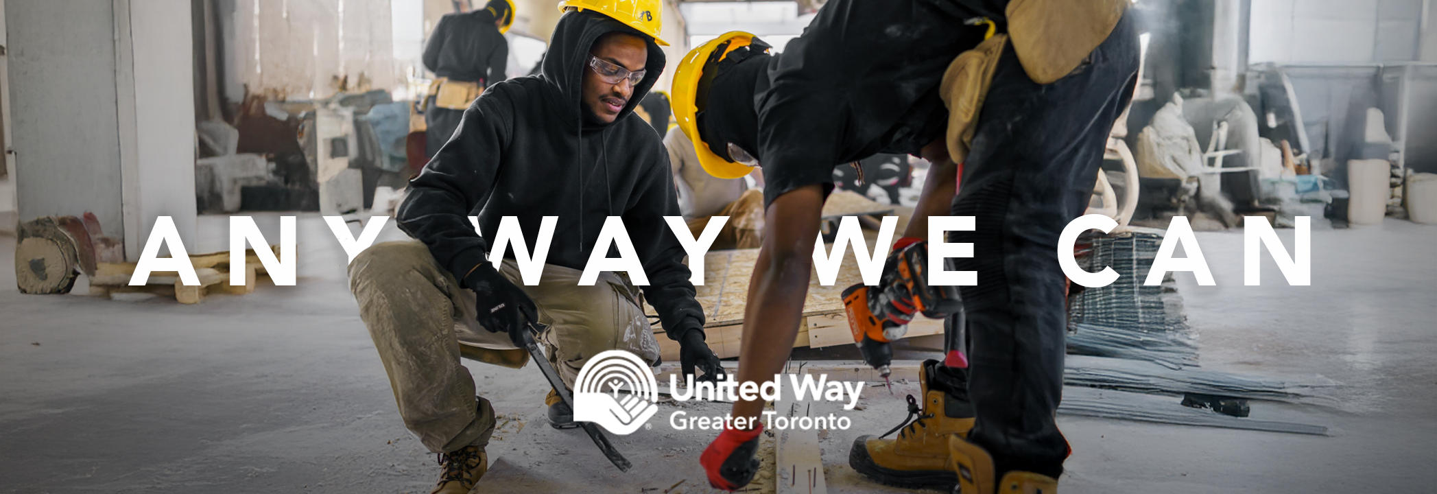 A Black man holds a saw while another Black man holds a piece of wood. Both wear hard hats in a building under construction. The words “Any way we can” and the United Way Greater Toronto logo are on top of the image in white.