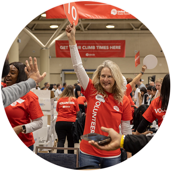 A white woman with gray hair wearing a red volunteer t-shirt holds up a sign in a packed room filled with volunteers.