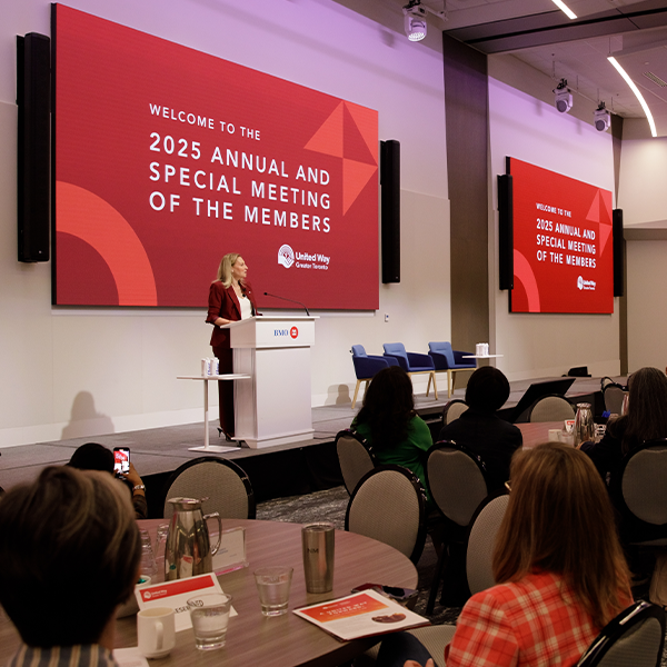 A person speaks at a podium in front of a large audience with a sign behind them that says “Welcome to the 2025 Annual and Special Meeting of the Members”