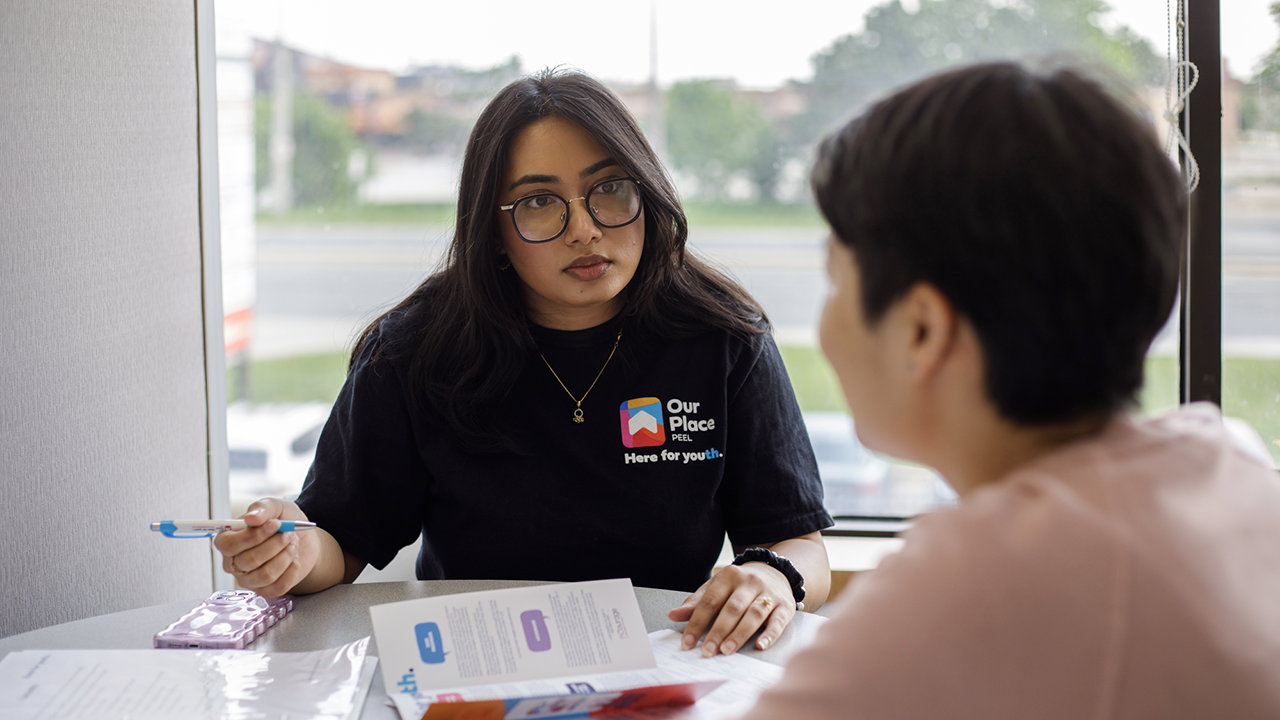 Hayah, an Intensive Case Manager at Our Place Peel, has long, dark brown hair and black-rimmed glassed. She wears a black t-shirt and sits at a desk holding a pen in her hand. She is talking to a person facing away from the camera with short dark hair.