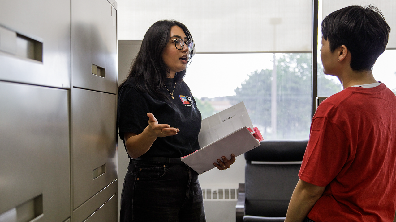 Hayah, an Intensive Case Manager at Our Place Peel, has long, dark brown hair and black-rimmed glassed. She wears a black t-shirt and pants. She is standing next to black filing cabinets talking to a person with short, dark hair and wearing a red t-shirt.