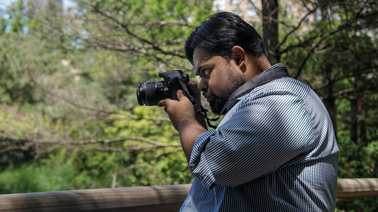 Zafeer looks through the viewfinder on his camera. He has brown skin, black hair and a black beard. He is wearing a blue and white striped shirt and has a camera around his neck. He is surrounded by lush, green trees on a sunny day.