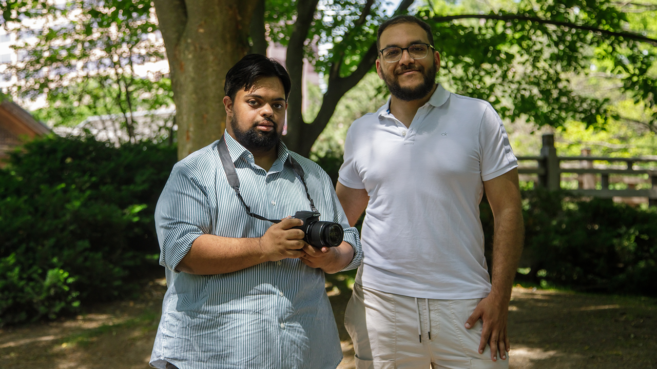Two men stand next to each other surrounded by green bushed and trees on a sunny day. The first has brown skin, black hair and a black beard. He is wearing a blue and white striped shirt, black pants and has a camera around his neck. The other is wearing a white shirt, khaki pants and glasses. He has brown skin, dark hair and a beard.