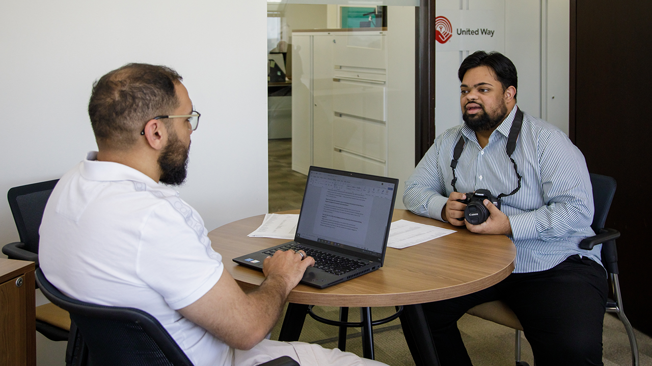 Two men sit in an office talking at a small, round table with papers scattered on top. One has his back facing the camera, is wearing a white shirt and khaki pants and glasses, has brown skin, dark hair and a beard and is typing on a laptop. The other has brown skin and black hair and beard, and is wearing a blue and white striped shirt, black pants and has a camera around his neck.