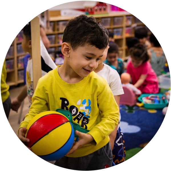 A child in a yellow shirt holds a toy ball in a playroom filled with other kids at Thorncliffe Park Community Hub. 