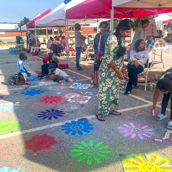 Community members gather under awnings at the launch of one of the 8 80 TowerPOPS, a project turning underused spaces in high-rise neighbourhoods into vibrant gathering places in Cooksville, Mississauga and Rockcliffe-Smythe, Toronto. Several youth are creating bright chalk images on the tarmac.