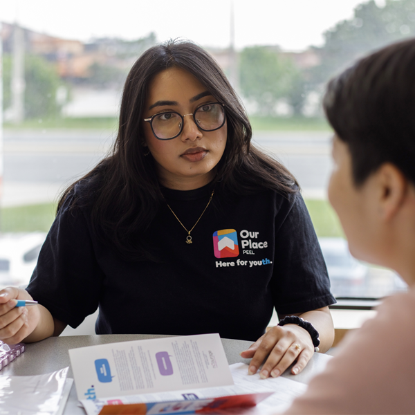 Hayah, a caseworker at United Way funded Our Place Peel, has long dark brown hair, black rimmed glasses and a blue shirt with an Our Place Peel logo on it. She sits at a table covered in papers talking to someone across the table who has short, black hair and a peach shirt.