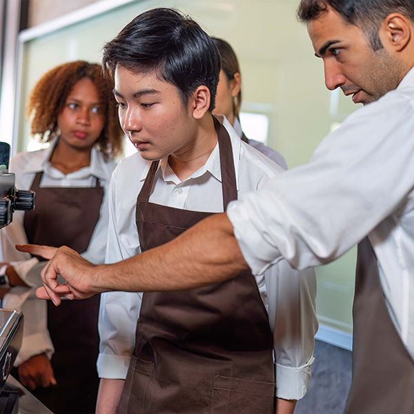  Several youth wearing white shirts and brown aprons gather around an industrial coffee maker as a teacher gives instructions.