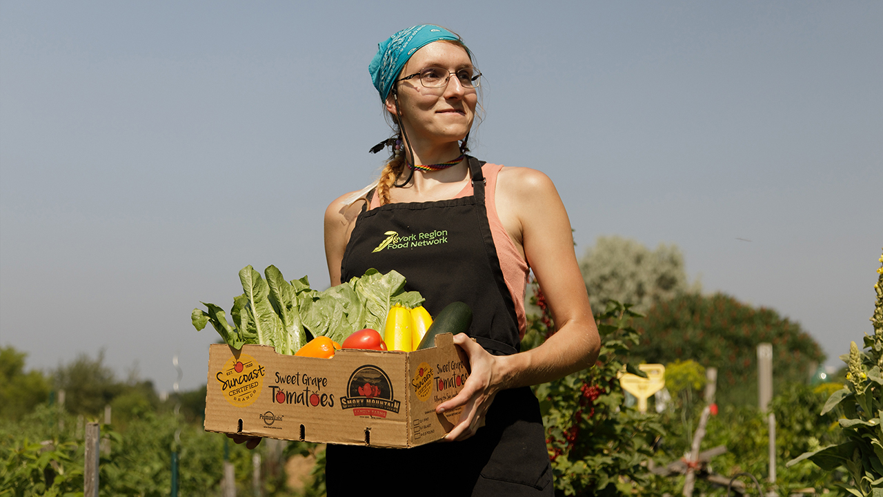 Anna stands outside on a bright summer day holding a box of produce from a United Way-funded Seeds of Change community garden program.