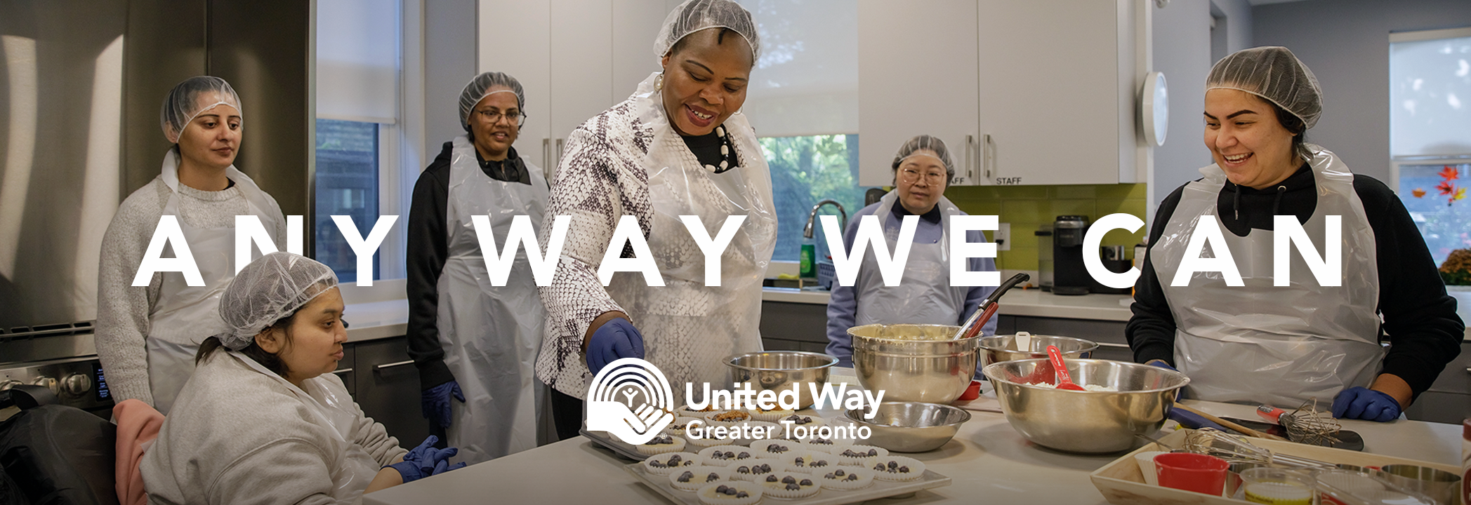 Six women wearing hairnets and aprons standing in a kitchen making desserts. 