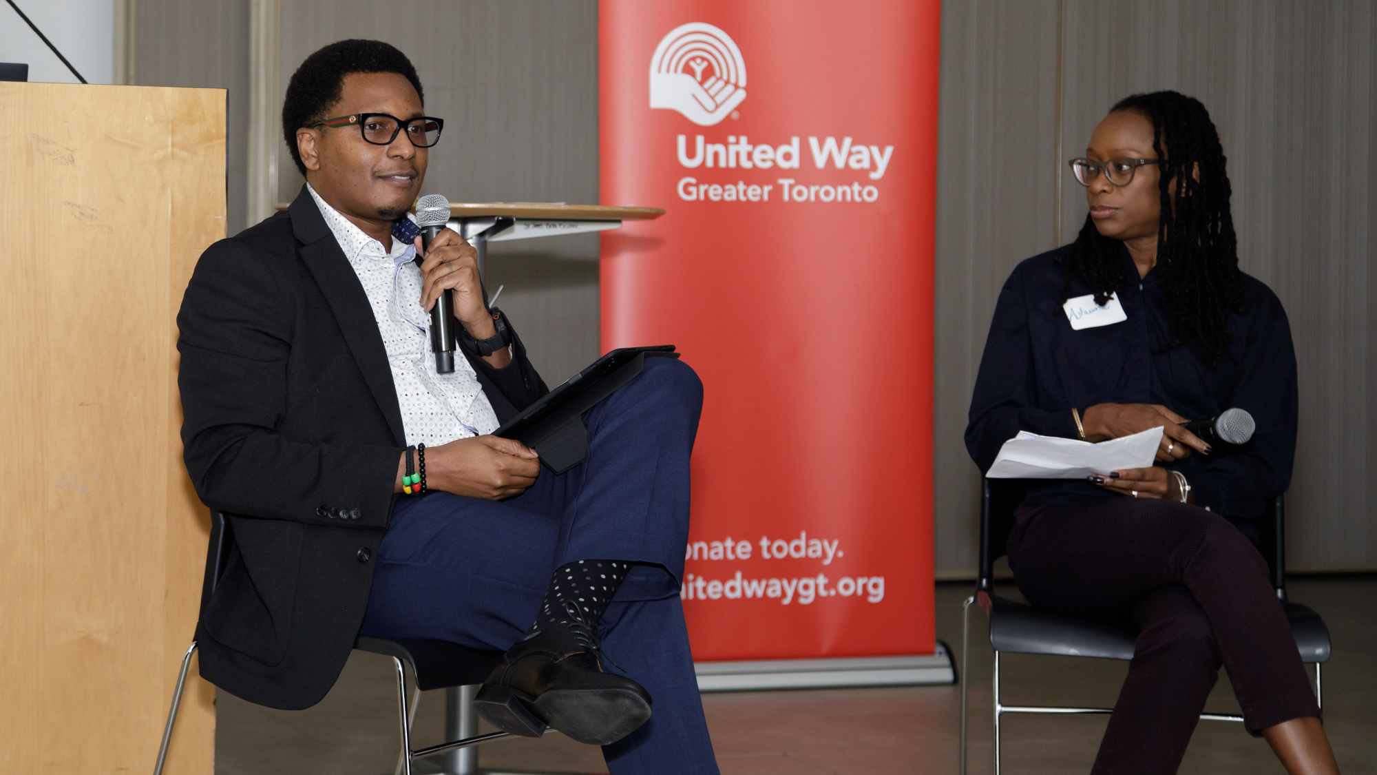 Jeff Msangi, Programs Manager at African Community Services of Peel, is a Black man wearing Black rimmed glasses, a white button up shirt, black suit jacket, blue pants and black shoes. He is speaking into a microphone at a United Way all-staff meeting and sitting across from Adaoma Patterson, Director of Community Service Investments at United Way Greater Toronto, a Black woman wearing brown-rimmed glasses, a dark blue button up shirt and black pants. Behind them is a banner with the United Way Greater Toronto logo. 