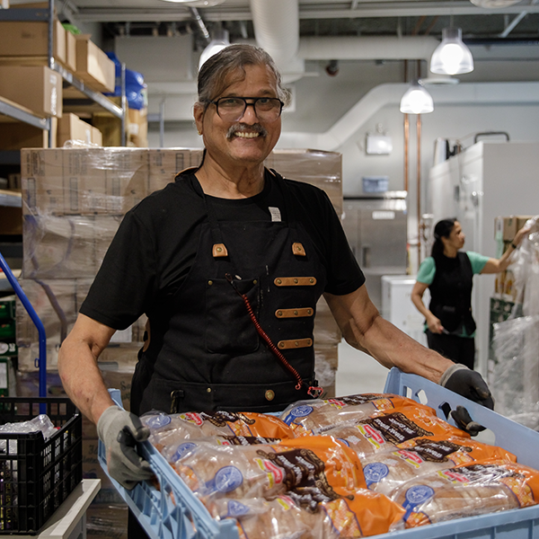 A person smiles at the camera and holds a pallet filled with bread at a local food bank. 