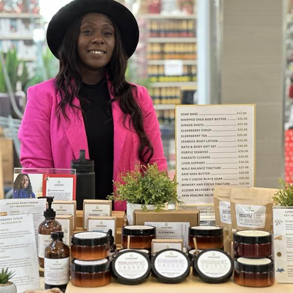 A Black woman wearing a black hat, black shirt and bright pink jacket, stands behind a table filled with products at the ILEO Holiday Market in November.