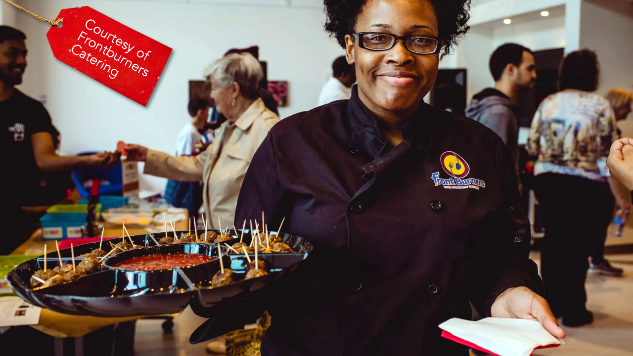Black woman with glasses wearing a Frontburners uniform holding a tray of appetizers at a function with a red gift tag icon with white text that says "Courtesy of Frontburners Catering"