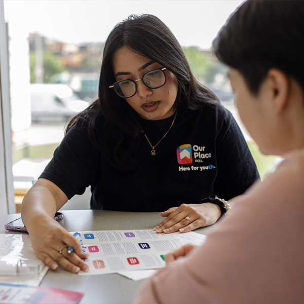 Young woman wearing an Our Place Peel t-shirt shows a piece of paper on a desk to another person