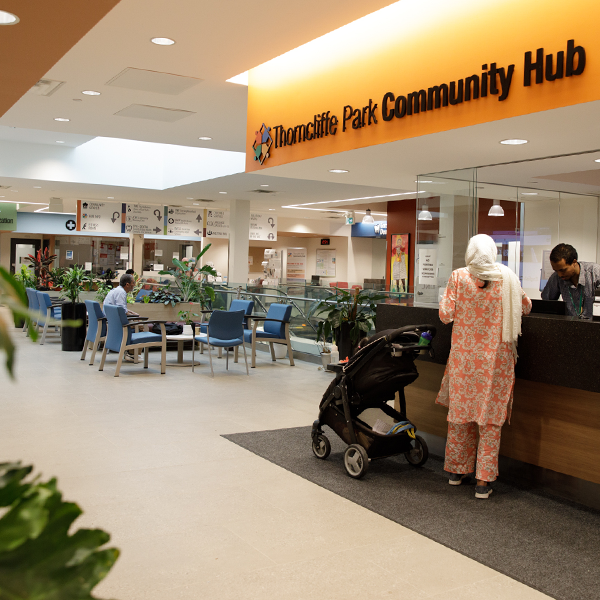 Woman with a stroller stands under a sign for Thorncliffe Park Community Hub, with people seated at tables in the background