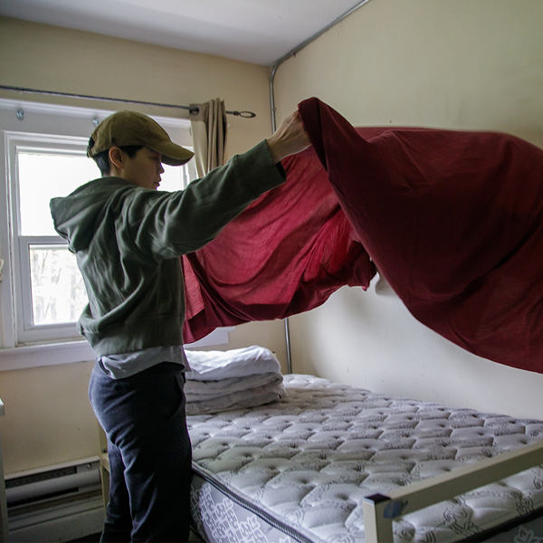 Young person wearing a cap holds a fitted sheet over a bare mattress