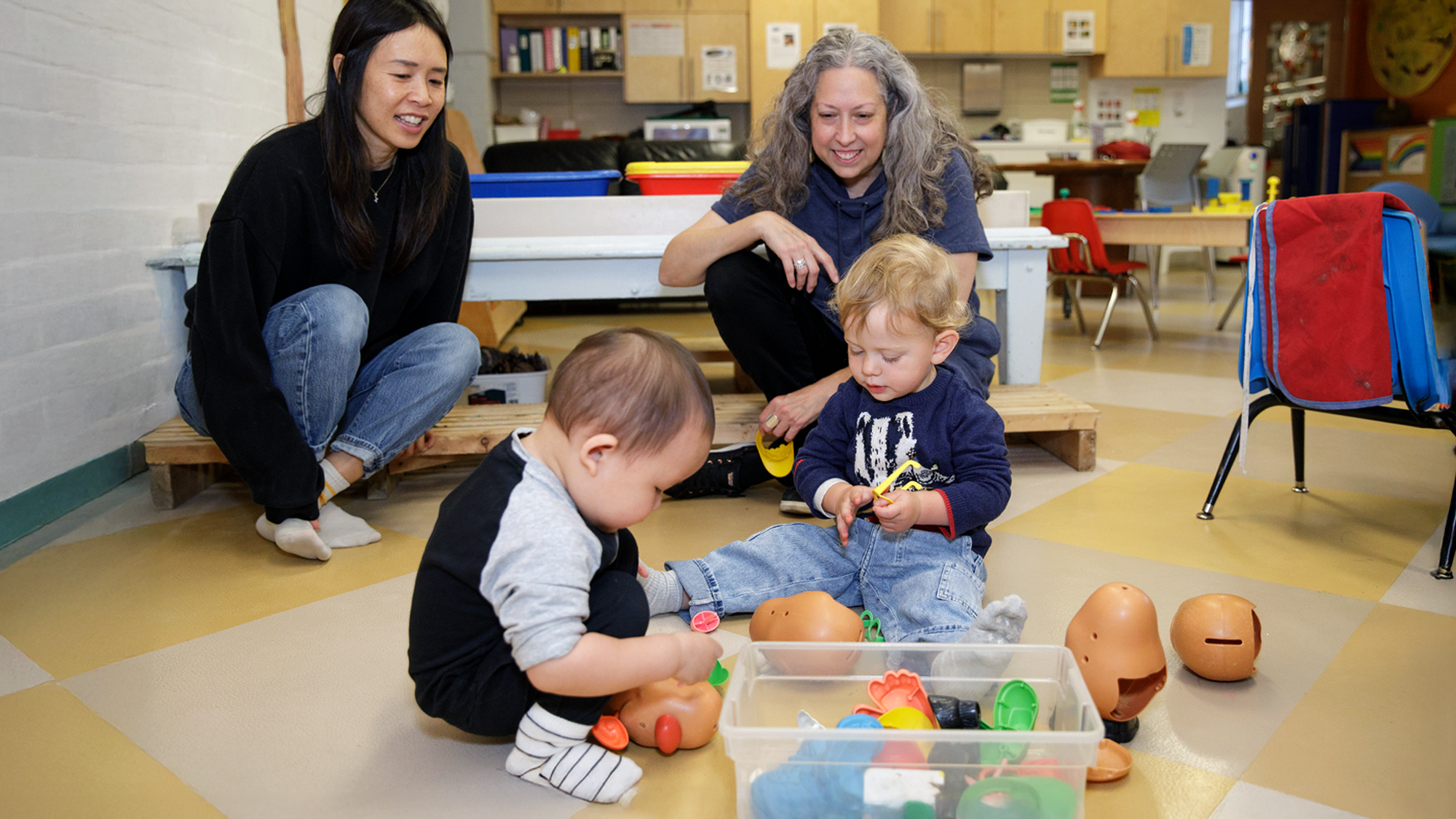 Two women watch two children playing with toys on the ground at College-Montrose Children’s Place