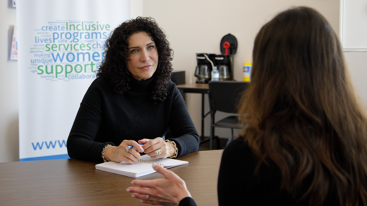 Piera, a light-skinned woman with dark curly hair and wearing a black turtleneck, sits at a desk listening intently to the woman sitting across from her At Women's Centre of York Region