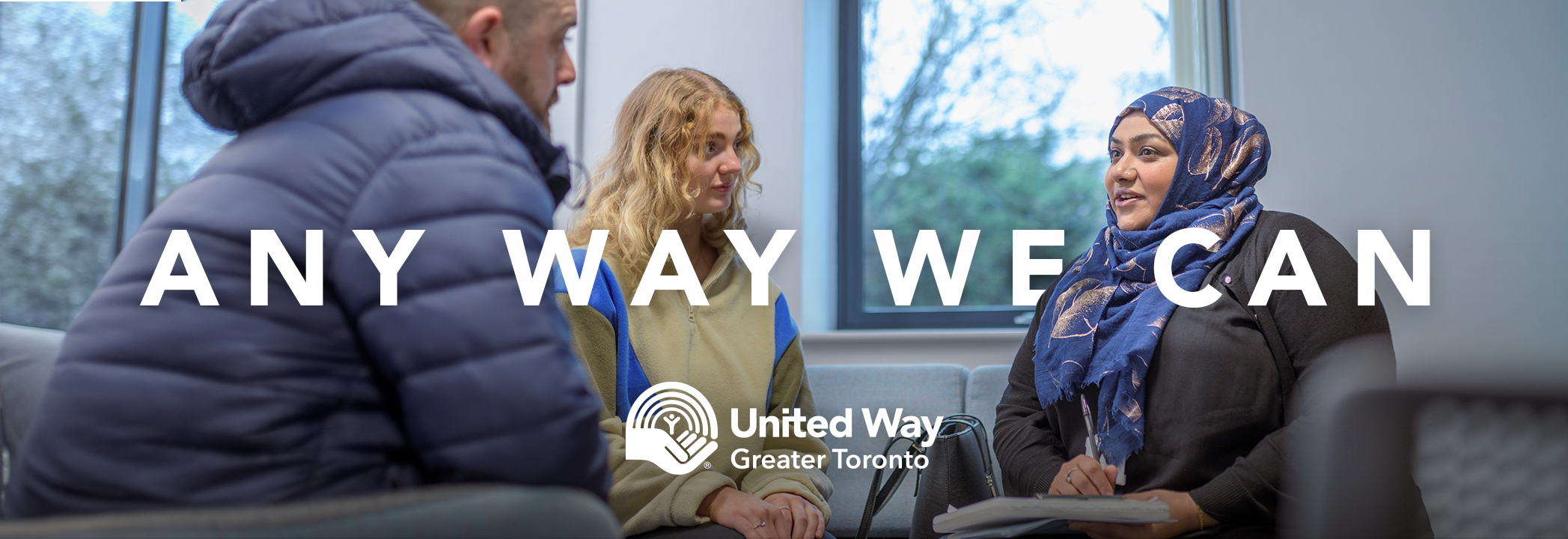 A woman writes in a notebook while speaking with two people seated across from her behind white text that says any way we can.