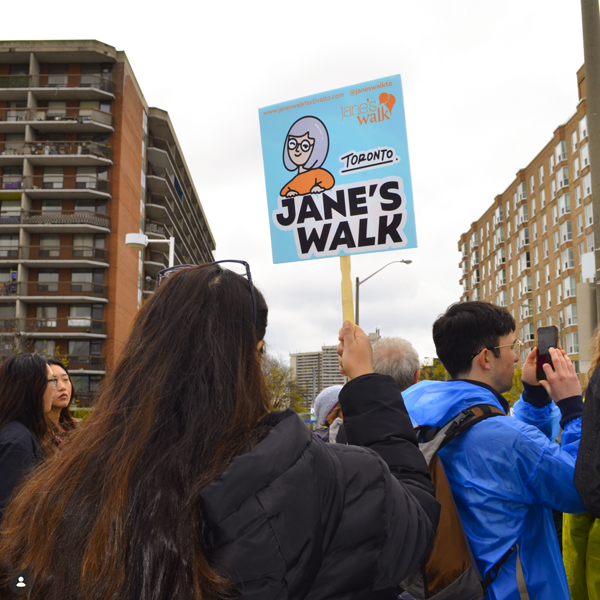 Participants walk together on a United Way neighbourhood tour, viewed from behind. One participant holds a sign that reads “Jane’s Walk.”