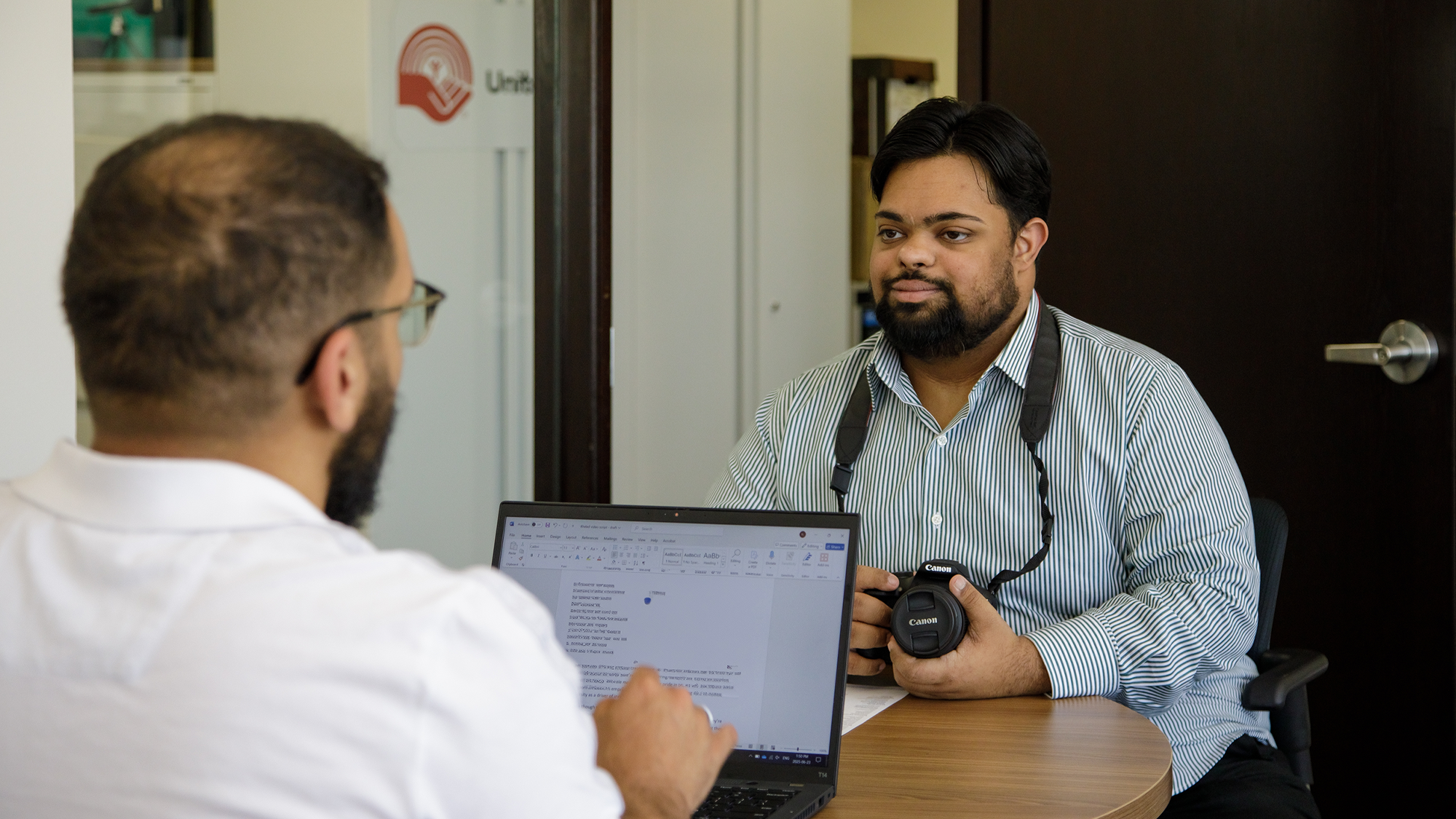 Two men sit at a small round table in an office, talking over papers and a laptop. One wears a white shirt and glasses and types on the laptop, while the other wears a blue striped shirt and has a camera around his neck.