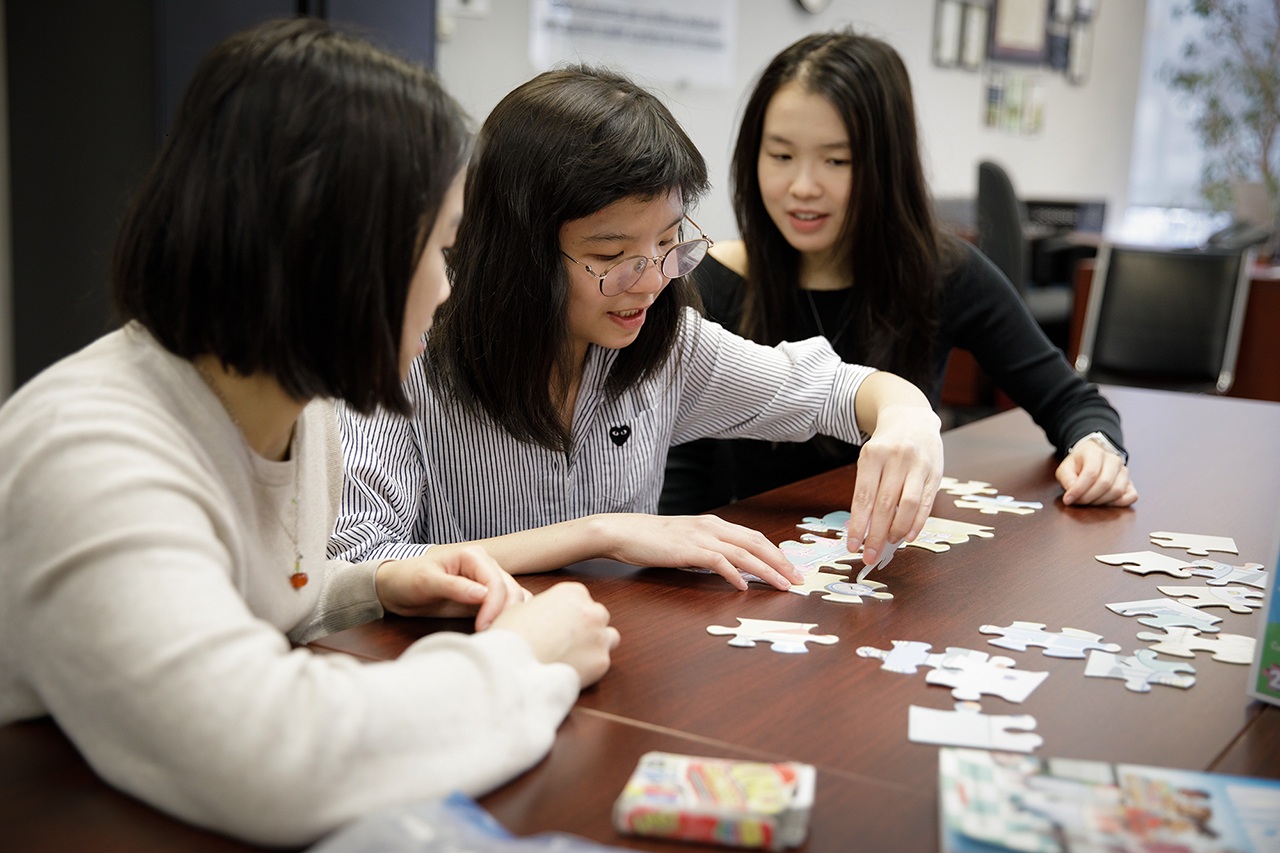 Bernice bends over a puzzle at a wooden table while sitting between her two sisters, smiling while focused on fitting the pieces together.