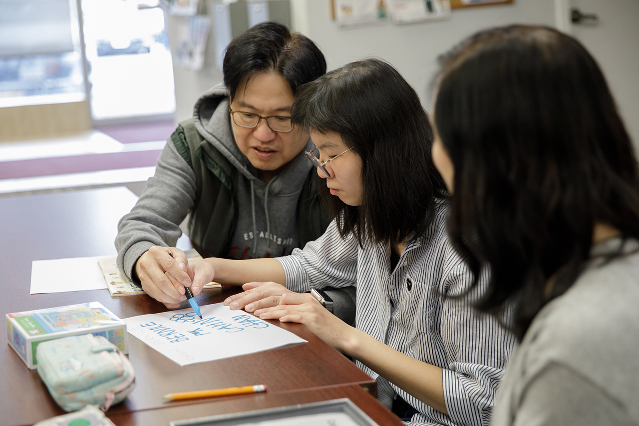 Bernice writes her name in blue marker on white paper while seated at a wooden table with her father, Ivan, helping beside her while her mother sits on her other side.