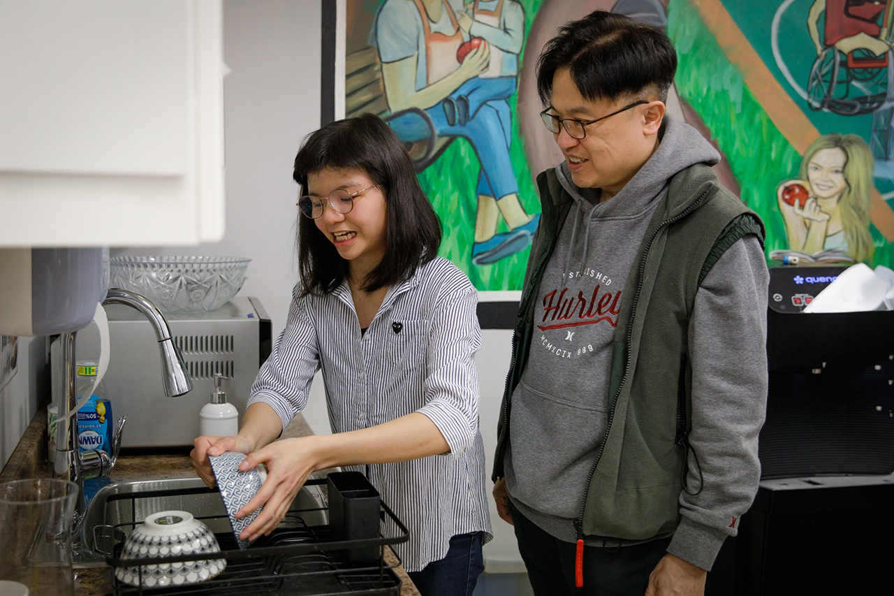 Ivan smiles as he stands behind his daughter Bernice in a kitchen while she leans over the sink washing a bowl.