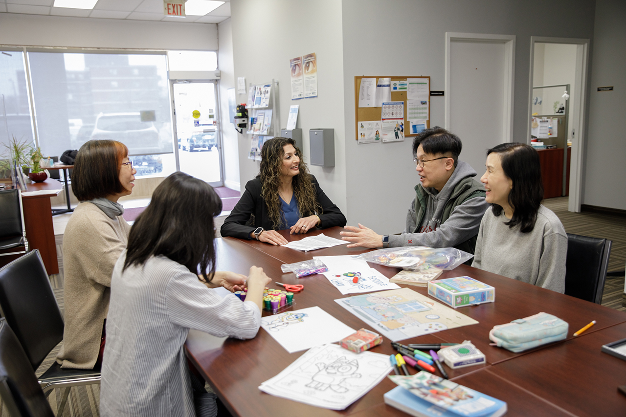 Bernice sits with her parents, Ivan and Annie, around a wooden table in a bright office meeting room. They are speaking with two staff members at Community Living York South. The table is covered with art supplies — markers, crayons, scissors, colouring pages and papers — suggesting a creative or educational planning session.