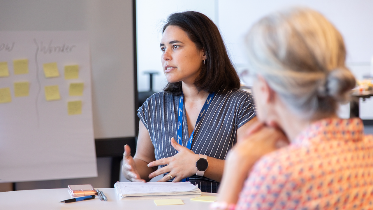 A woman sits at table, gesturing with her hands while speaking to another woman, whose back is turned to the camera with her blonde hair in a bun. Behind them, there is a white board with multiple yellow sticky notes.