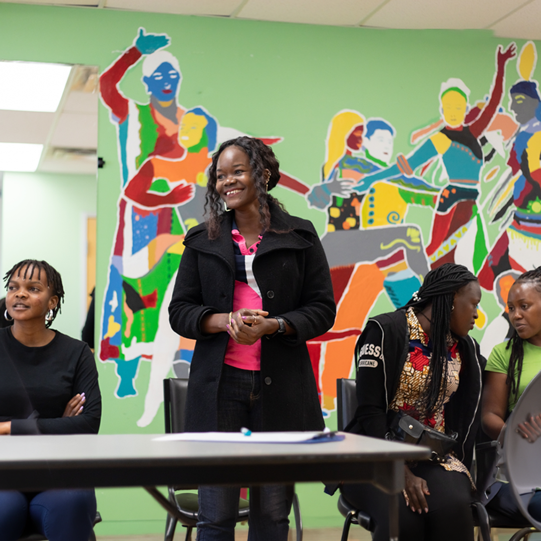 A woman is standing at table smiling in front of a colourful mural in a green room with people sitting around her. There is another woman sitting next to her at the table and two people to her right speaking to one another.