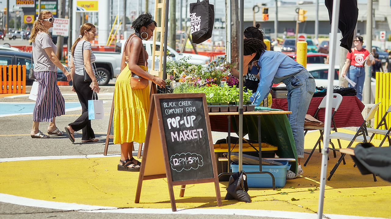 A woman in a yellow dress stands in front of a stand at the Black Creek Community Farm Pop-up Market.