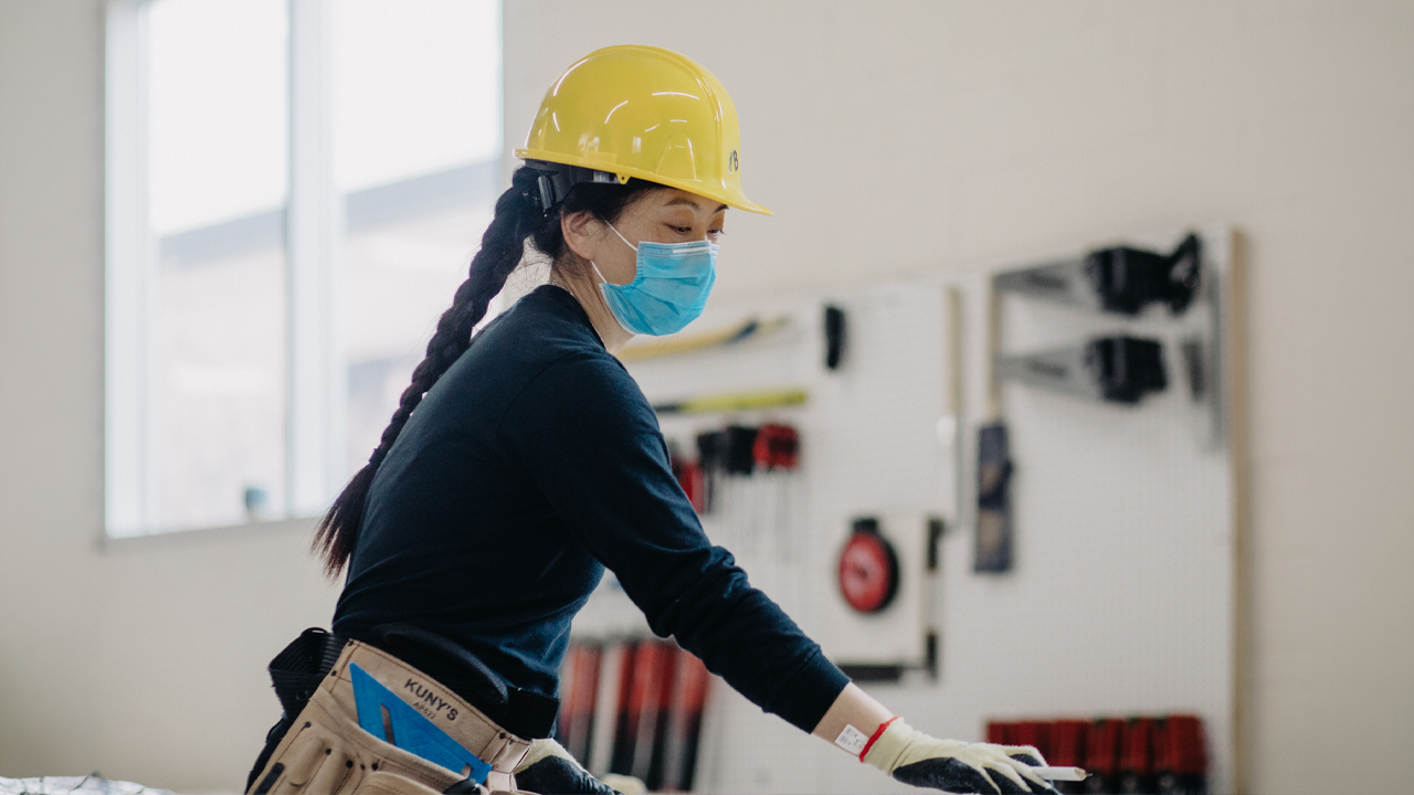 A woman wearing a hard hat, face mask and tool belt works on a construction project in a training workshop.