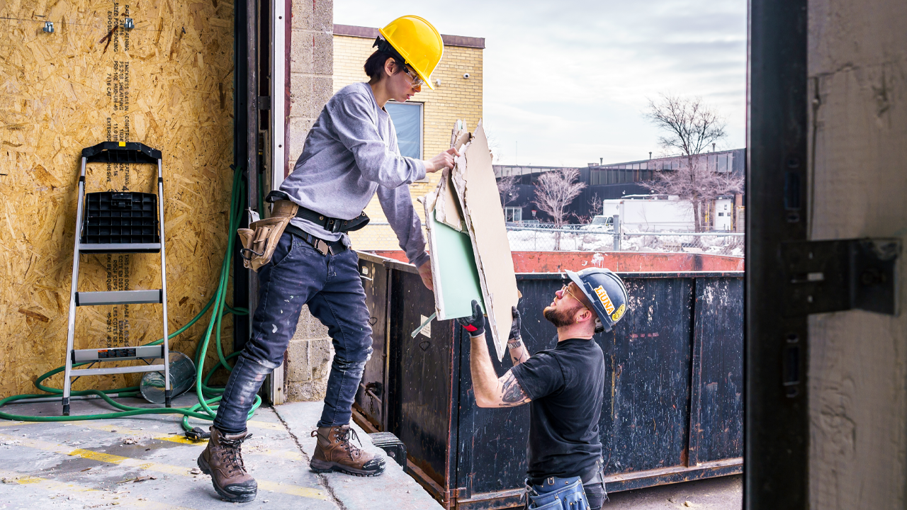 A worker in a hard hat passes a piece of drywall to a coworker.