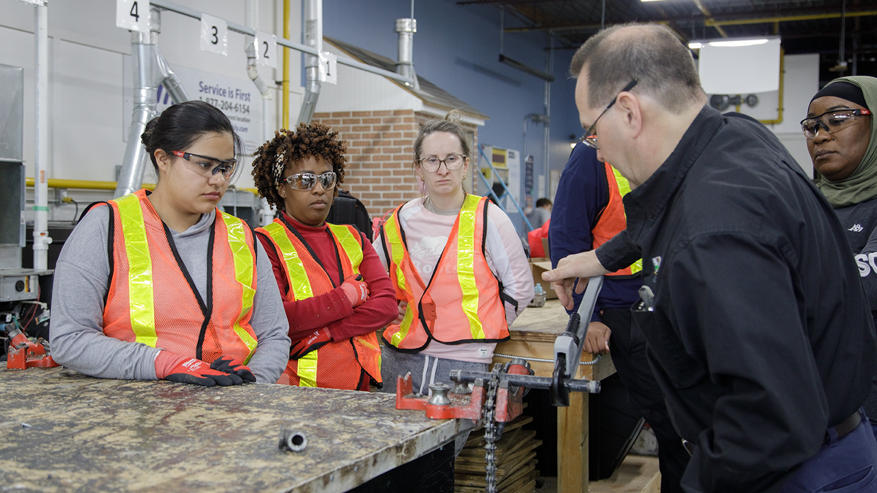 Participants in The Redwood's Women in Trades & Technology program, wearing reflective safety gear, gather around an instructor demonstrating a piece of equipment.