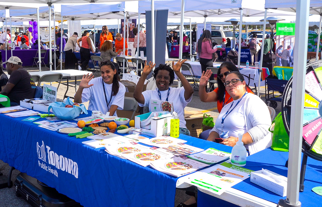 Volunteers wave to the camera while sitting behind a stall at the Community Wellness Fair.