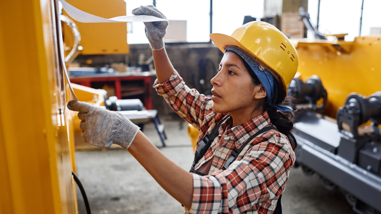 A woman wearing a yellow hard hat, gloves and a plaid shirt references a document on a work site. 
