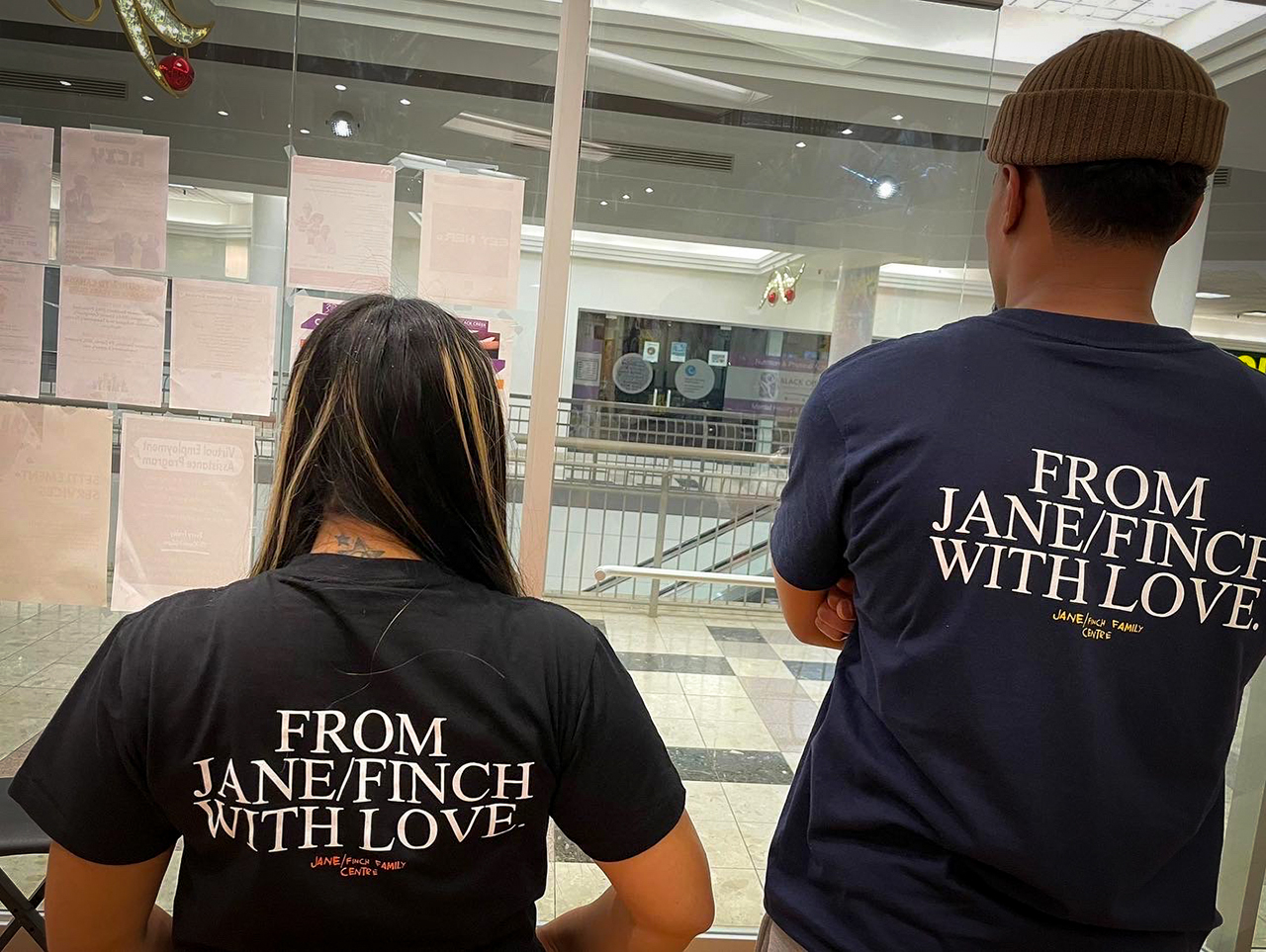 Two people face away from the camera wearing black T-shirts that have "From Jane/Finch with love" written on them in white text.